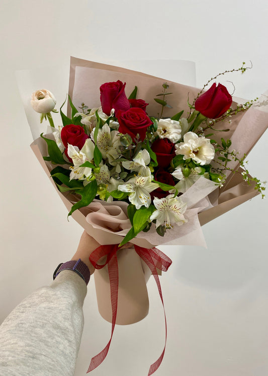 Hand-tied bouquet featuring red roses and white seasonal flowers, wrapped in soft pink paper with a red ribbon.