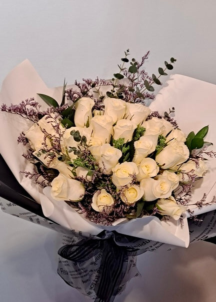 A hand-tied bouquet of 36 white roses accented with greenery and delicate fillers, wrapped in elegant black and white paper with a ribbon.