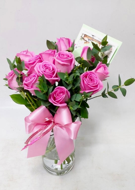 A floral arrangement of 12 fresh pink roses in a clear glass vase, decorated with greenery and a pink satin ribbon.