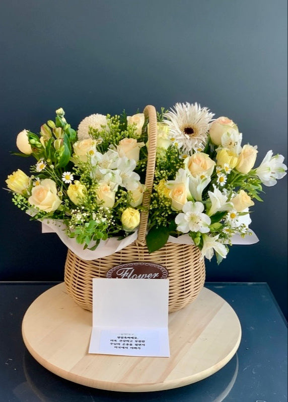 Pastel-toned flower basket arranged with yellow and peach blooms, presented in a woven basket with lush greenery.
