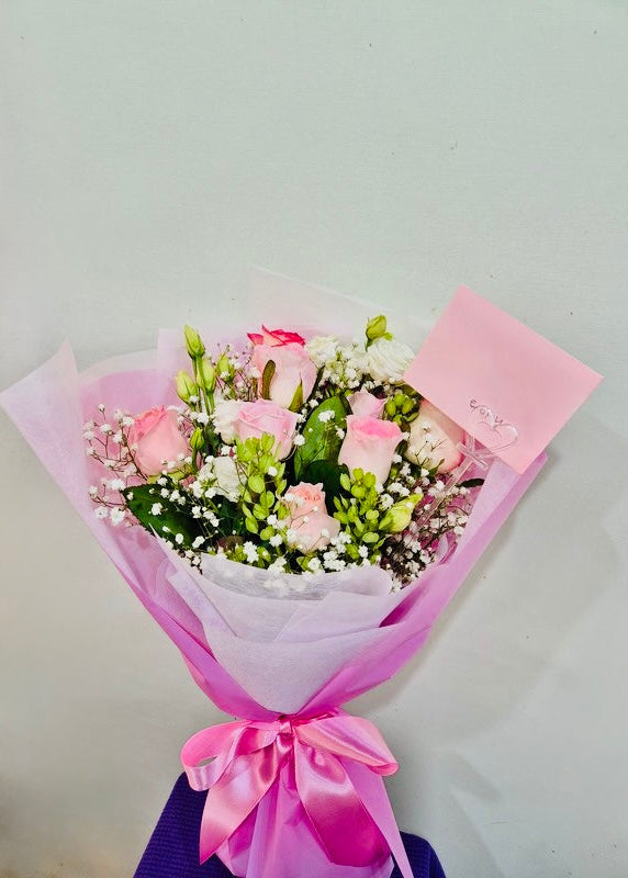 Hand-tied bouquet of seven pastel pink roses with white lisianthus, baby’s breath, and greenery, wrapped in soft pink paper with a ribbon, perfect for a romantic or thoughtful gift