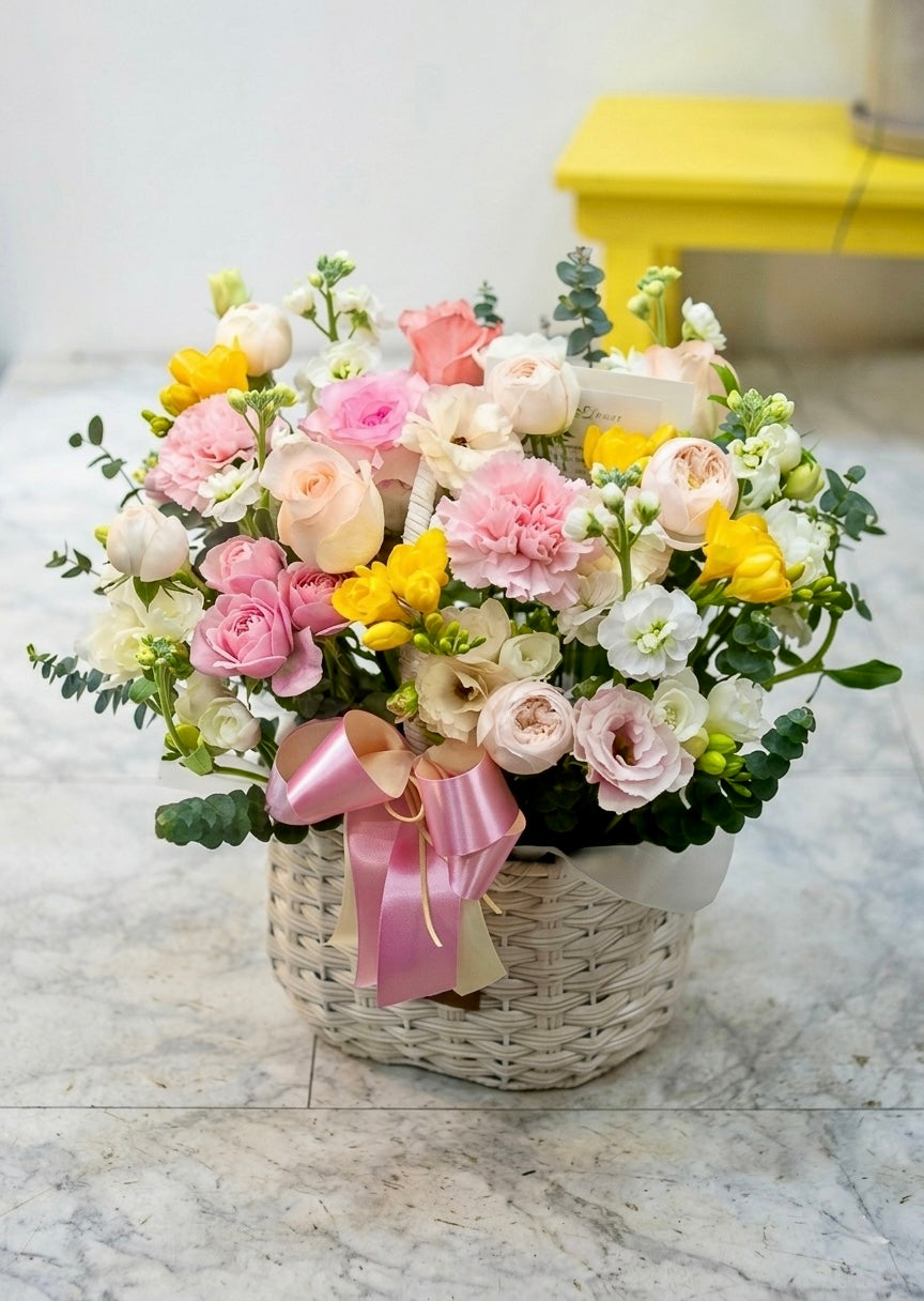 Pastel flower basket arrangement featuring pink, white, and yellow blooms with fresh greenery in a woven basket, perfect for birthdays, congratulations, and thoughtful gifts.