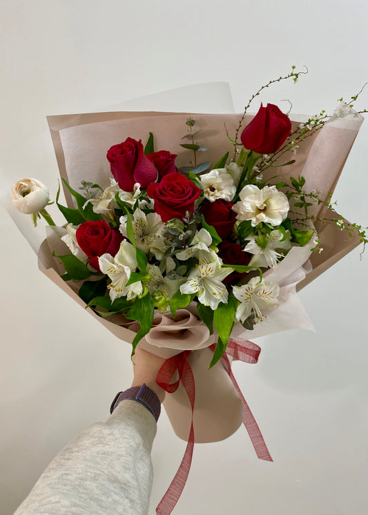 Hand-tied bouquet featuring red roses and white seasonal flowers, wrapped in soft pink paper with a red ribbon.