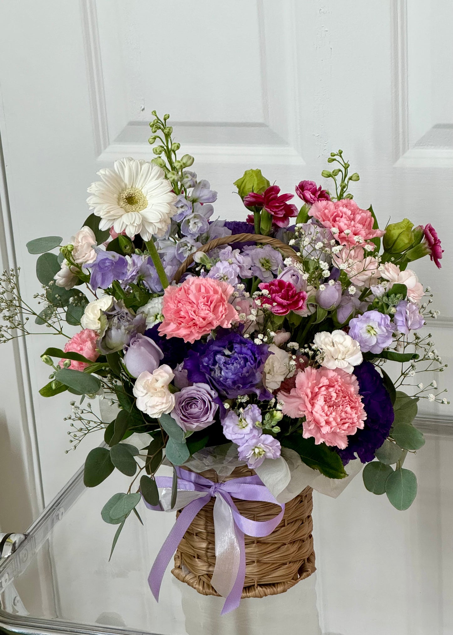 A cheerful mixed flower basket featuring pink carnations, purple lisianthus, lavender roses, and white gerbera daisies, arranged with greenery in a woven basket and finished with a lavender ribbon.