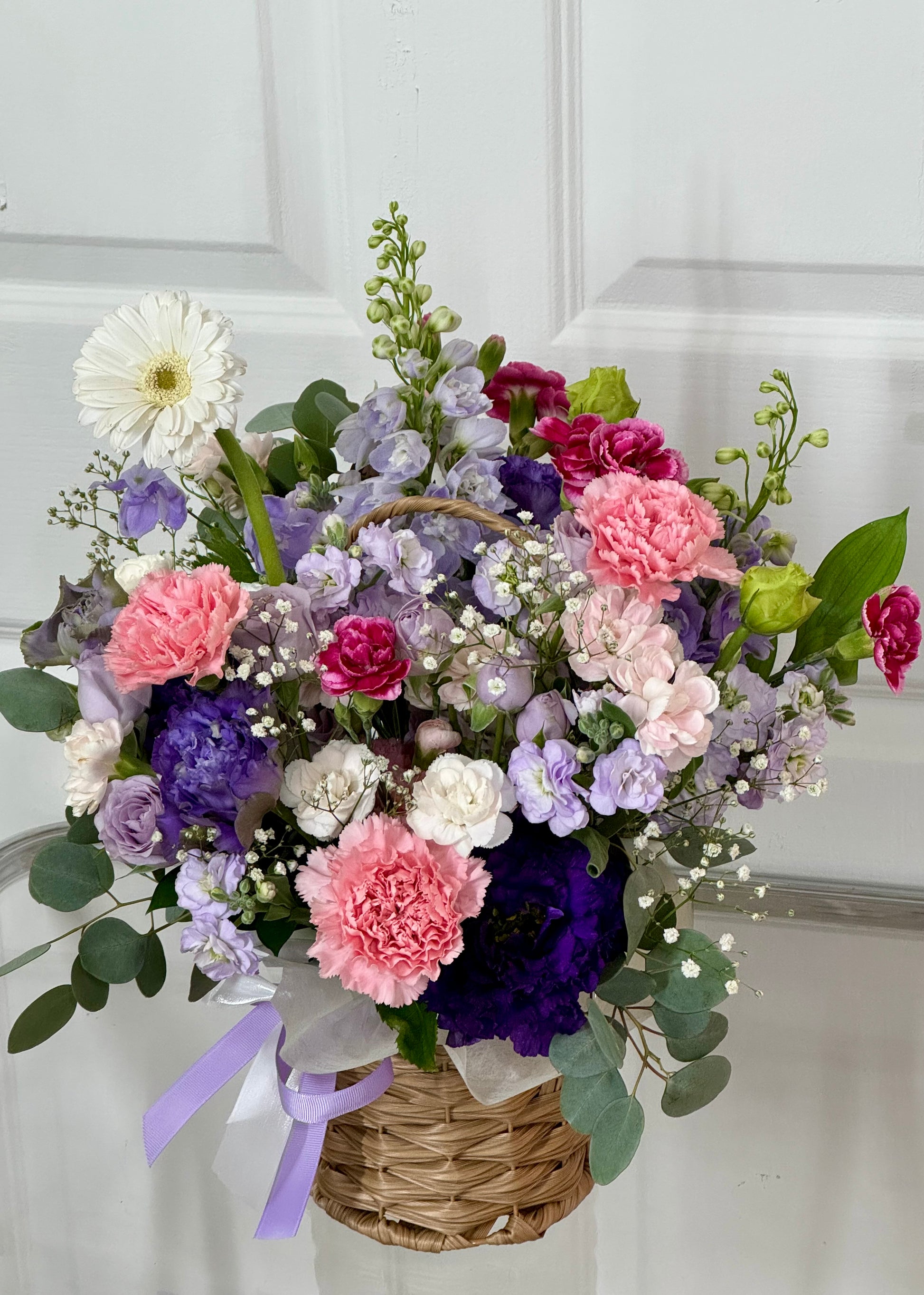 A cheerful mixed flower basket featuring pink carnations, purple lisianthus, lavender roses, and white gerbera daisies, arranged with greenery in a woven basket and finished with a lavender ribbon.