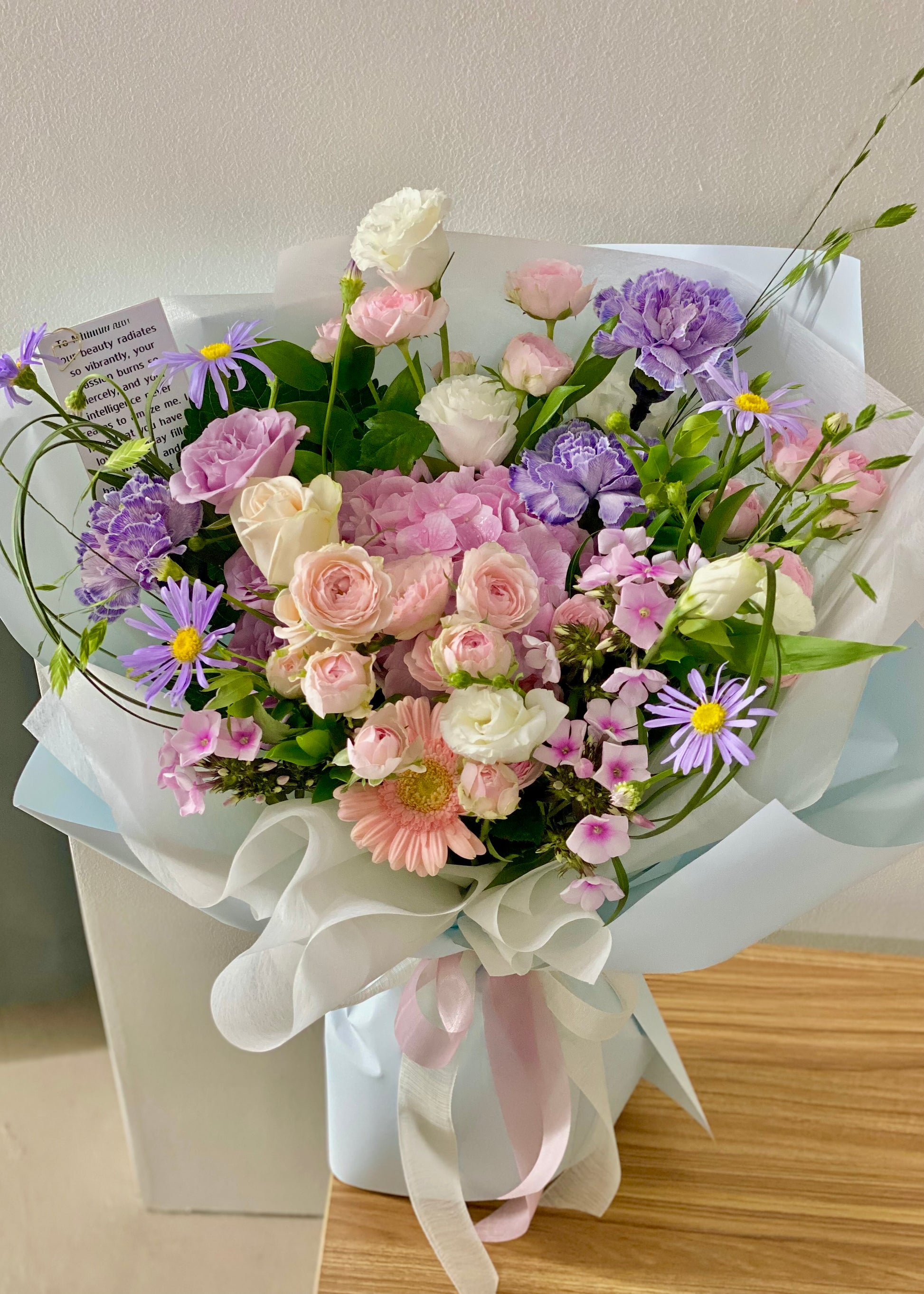 Hand-tied bouquet of soft pink and lavender flowers wrapped in pastel paper, ideal for sympathy, love, or everyday gifting.