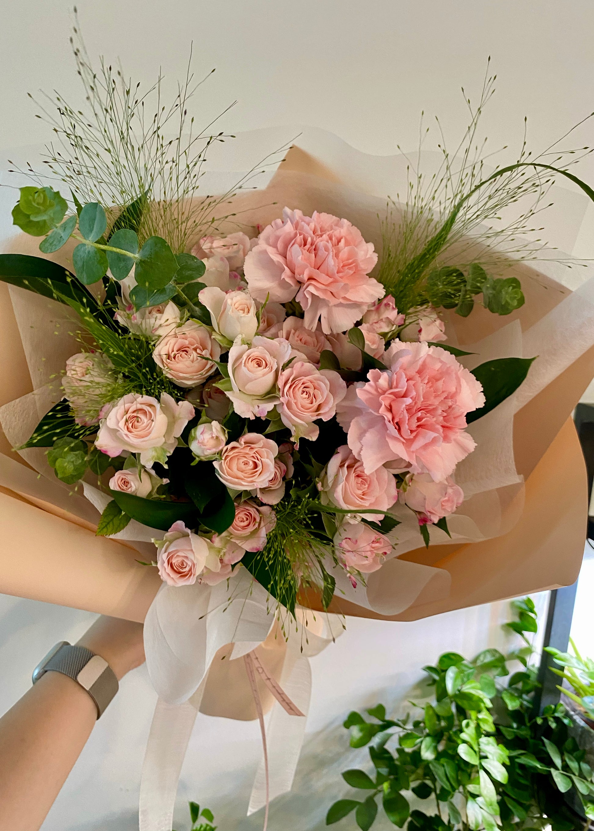 Hand-tied bouquet of soft pink roses and carnations, arranged with elegant greenery and wrapped in beige and white paper.