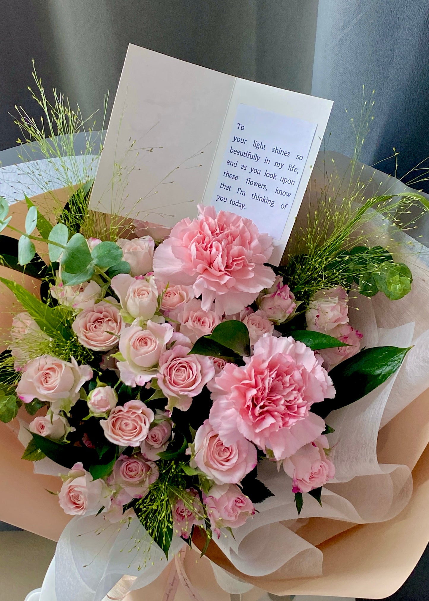 Hand-tied bouquet of soft pink roses and carnations, arranged with elegant greenery and wrapped in beige and white paper.