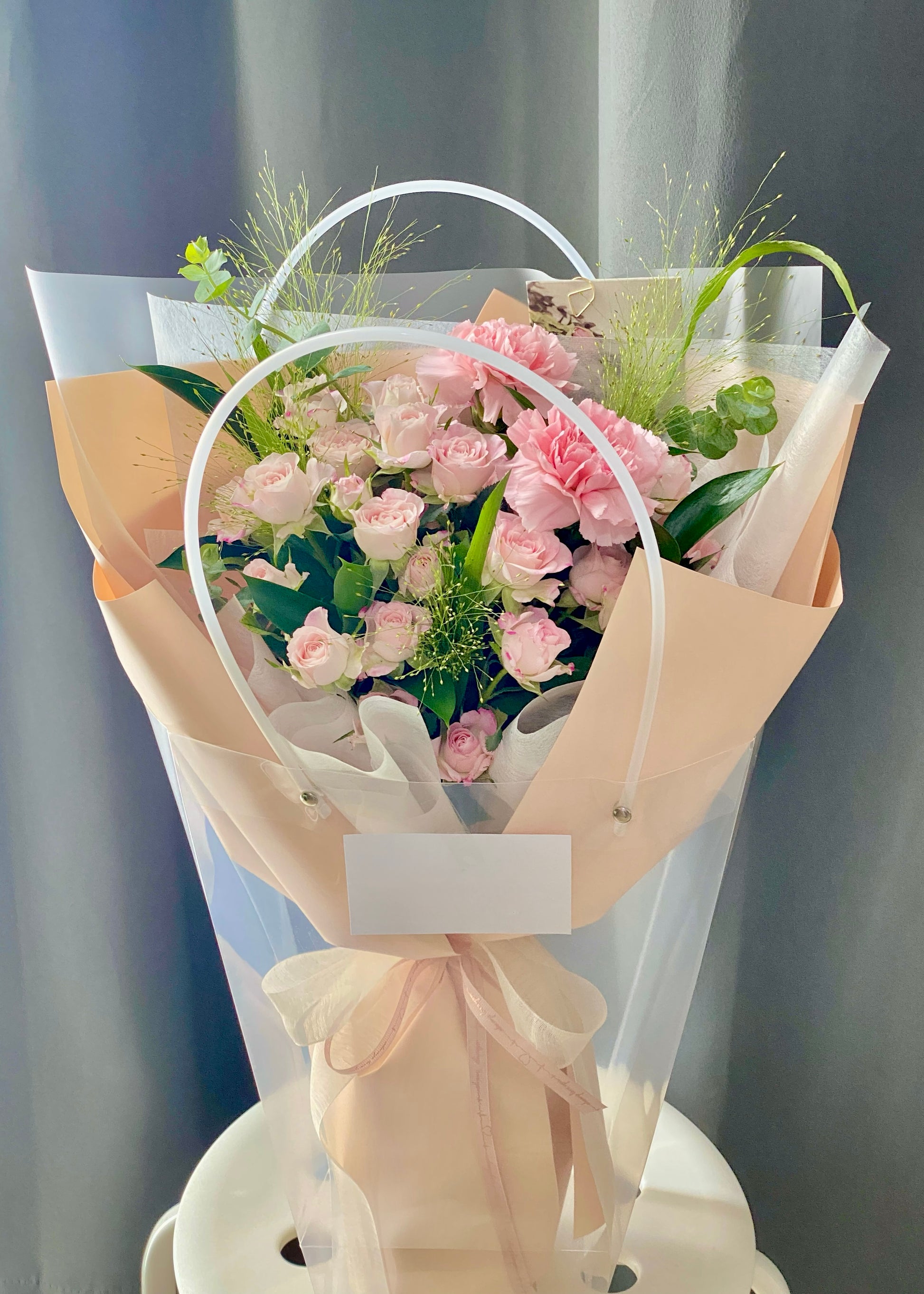 Hand-tied bouquet of soft pink roses and carnations, arranged with elegant greenery and wrapped in beige and white paper.