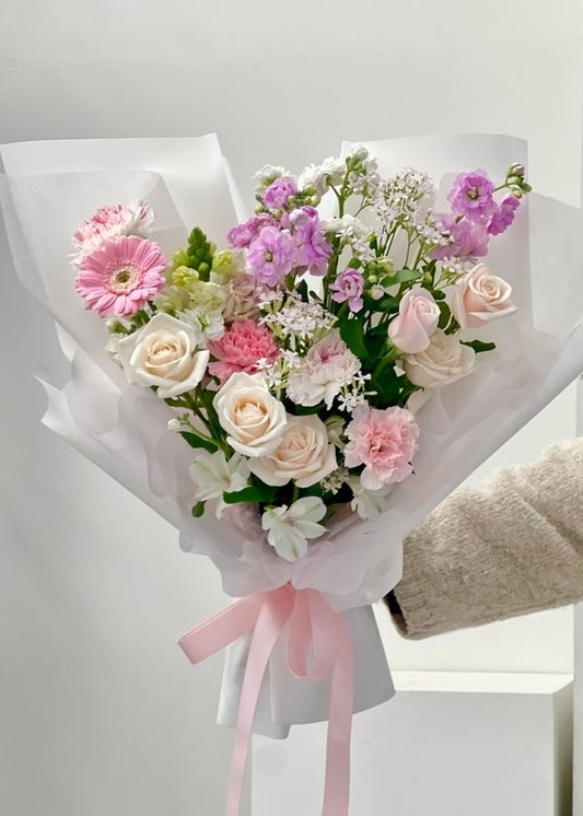 Hand-tied bouquet featuring pastel roses and mixed seasonal flowers in soft pink, lavender, and cream tones, wrapped in delicate paper with greenery accents.